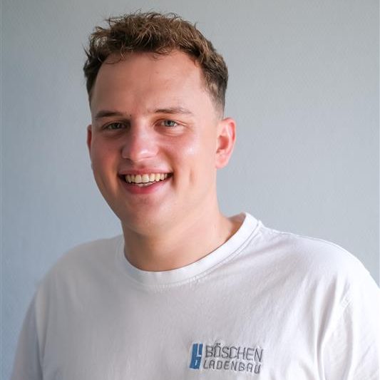 Porträt von Hendrik Böschen in weißen T-Shirt mit Bö­schen Ladenbau-Logo, vor hellem strukturiertem